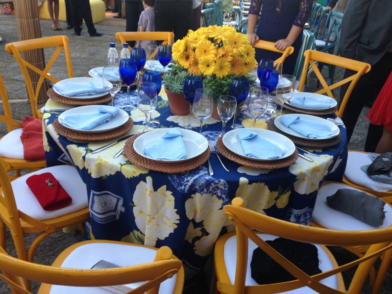 Os sous-plats em palhinha, a base da toalha da mesa em azul e as cadeiras amarelas ficaram ainda mais em evidencia com os copos azuis e arranjo com flores amarelas.