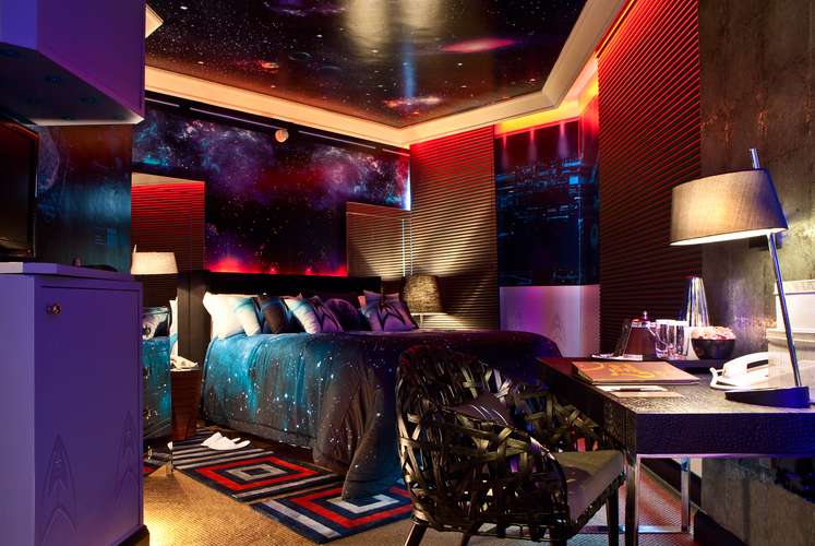 A cena se transforma totalmente quando a luz muda de tonalidade. Com o vermelho podemos perceber a cama com colha inspirada no infinito do espaço. E em primeiro plano a cadeira trançada mostra que é possível adaptar um quarto com elementos tradicionais.